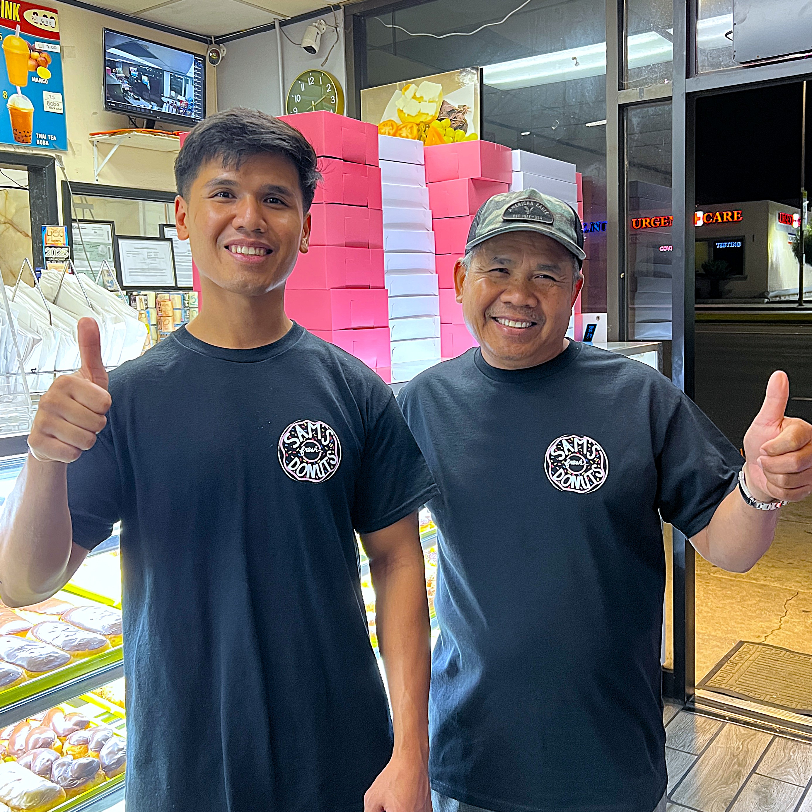 Sam and his son smiling behind the counter at Sam's Fresh Donuts, giving thumbs up in front of stacks of pink-and-white donut boxes.